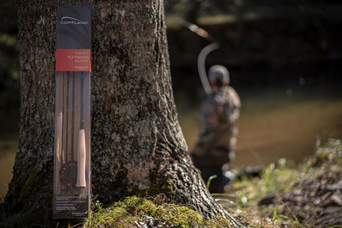 Fairplay Outfit in its packaging leaning against a tree. In the background someone is fly fishing