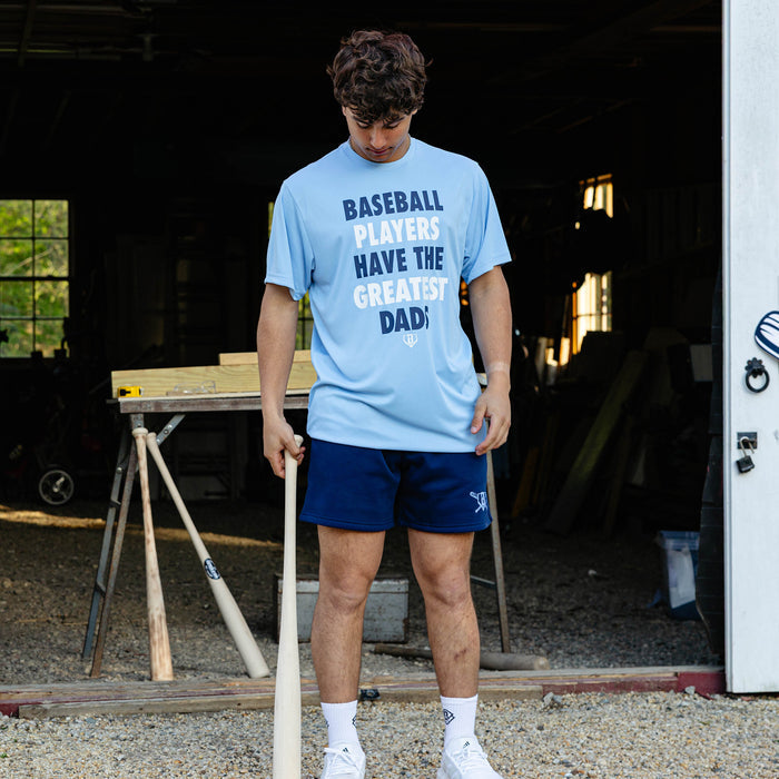 Young adult male model looking down while holding a wooden baseball bat and wearing the navy Heritage Diamond Sweatshort paired with the columbia blue Greatest Dad Athletic Tee and white BL101 crew socks.