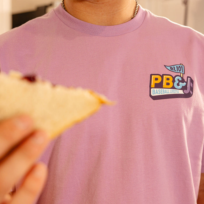 Close-up of the PB&J Baseball Lifestyle chest logo on the purple tee, with a PB&J sandwich in the foreground.