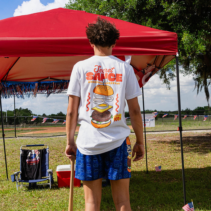 Person wearing a white secret sauce. t-shirt and blue Secret Sauce burger print shorts holding baseball bat under a red canopy at an outdoor event.