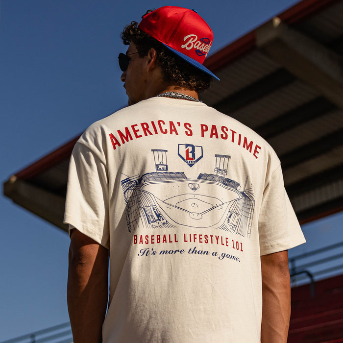 Back view of the BL101 America's Pastime cream tee on a male model at a stadium, showing the stadium sketch graphic and "It's more than a game" text.