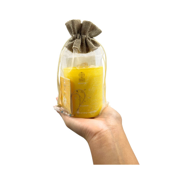 Hand holding a yellow candle in a glass jar with a brown drawstring bag on a white background