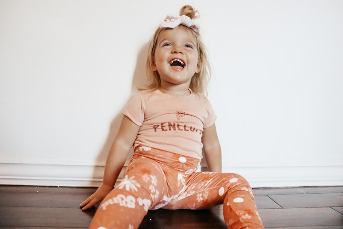 Child wearing a pink shirt with 'Penthouse' text and orange pants with white patterns, sitting on a wooden floor.