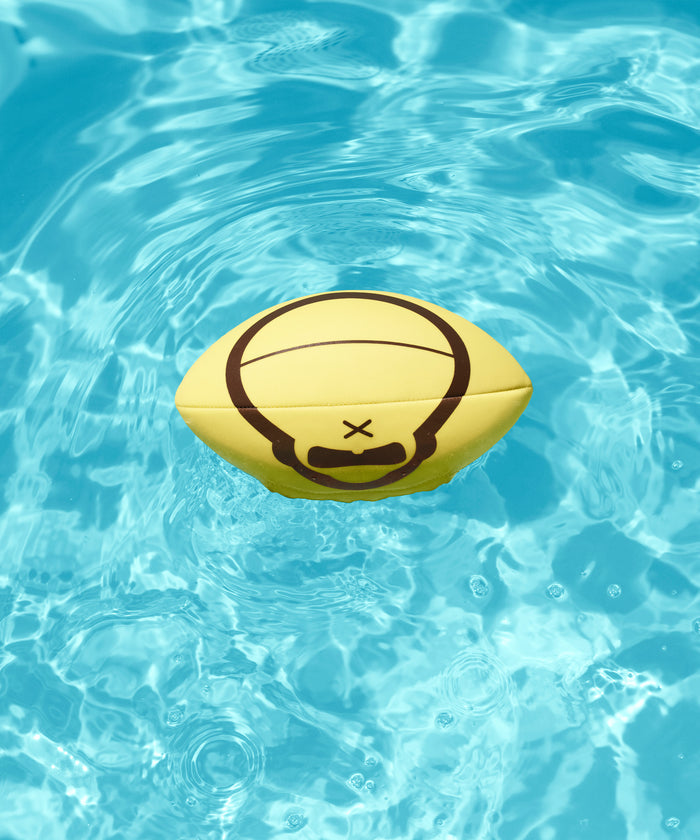 Yellow Beach Football with a Sun Bum logo floating on clear blue water in a pool setting.