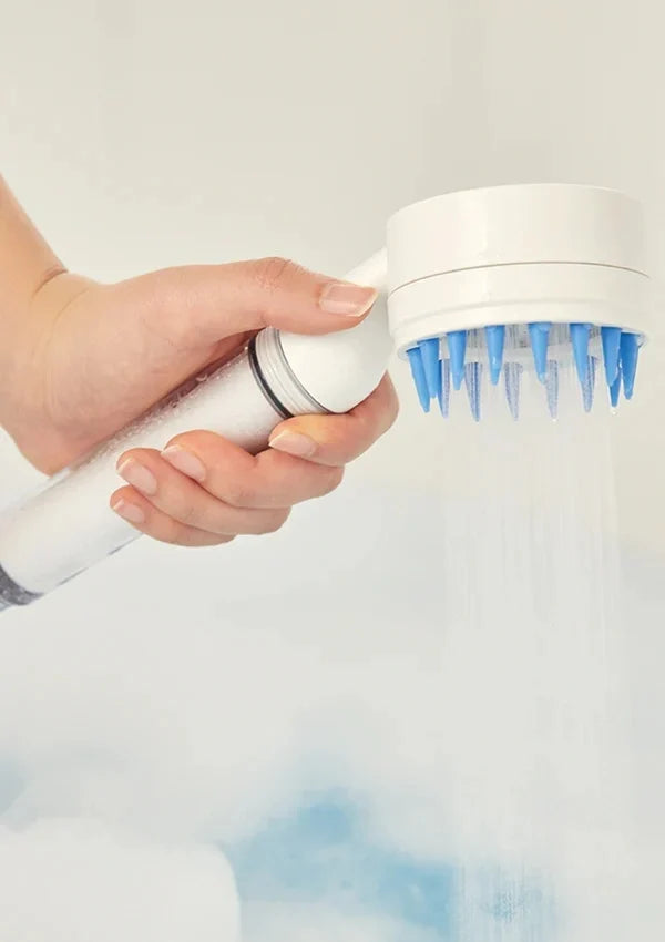 A hand holding a white handheld shower head with blue silicone spray nozzles, spraying water against a soft white background.