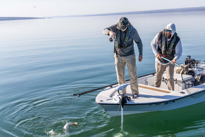 Two anglers are standing on a boat. One has prepared the net, while the other is reeling in the fish he caught.