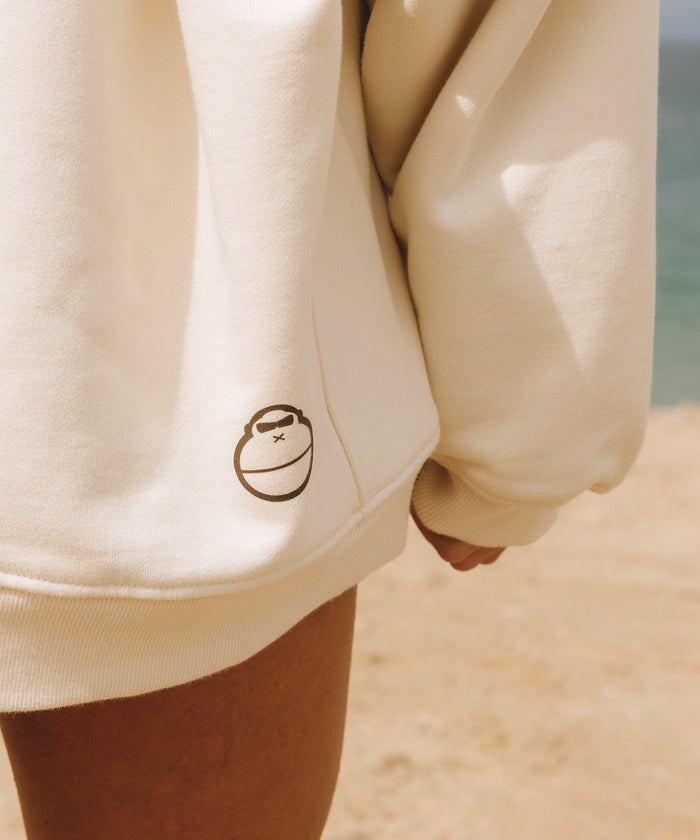 Close-up of a person wearing a cream-colored hoodie on a sandy beach, with the small black circular logo of the brand 'Kaleidoscope' visible on the lower left of the chest.