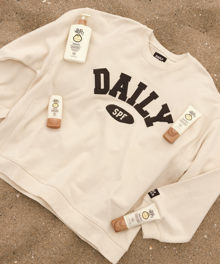 An off-white crewneck sweatshirt with 'DAILY SPF' printed in black across the chest, styled on a sandy surface with sunscreen bottles and tubes placed around it.