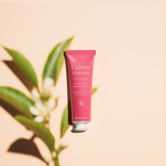 A pink tube of Manucurist Everyday Orange Blossom Hand Cream - Cosmos Organic stands upright on a beige background, featuring white text and coconut oil, while a leafy plant casts a soft shadow nearby.