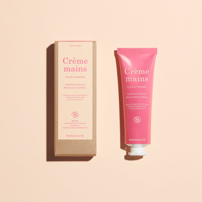 A pink tube of Manucurist Everyday Orange Blossom Hand Cream - Cosmos Organic sits beside its brown rectangular box on a light beige background, with both items displaying French text.