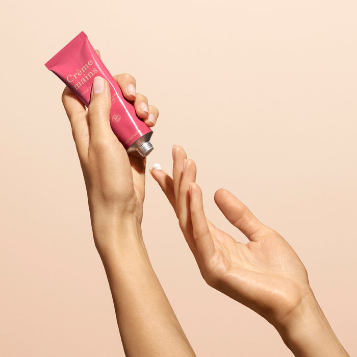 A person squeezes a small amount of Manucurist Everyday Orange Blossom Hand Cream - Cosmos Organic from a pink tube onto their finger against a beige background.