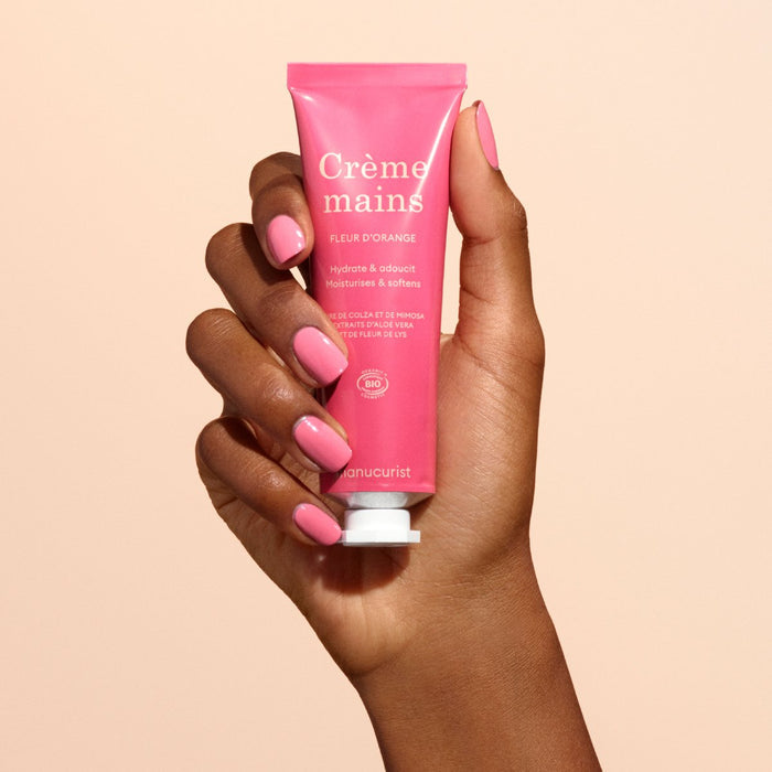 A hand with pink nail polish holds a pink tube of Manucurist Everyday Orange Blossom Hand Cream - Cosmos Organic, enriched with coconut oil, against a beige background.