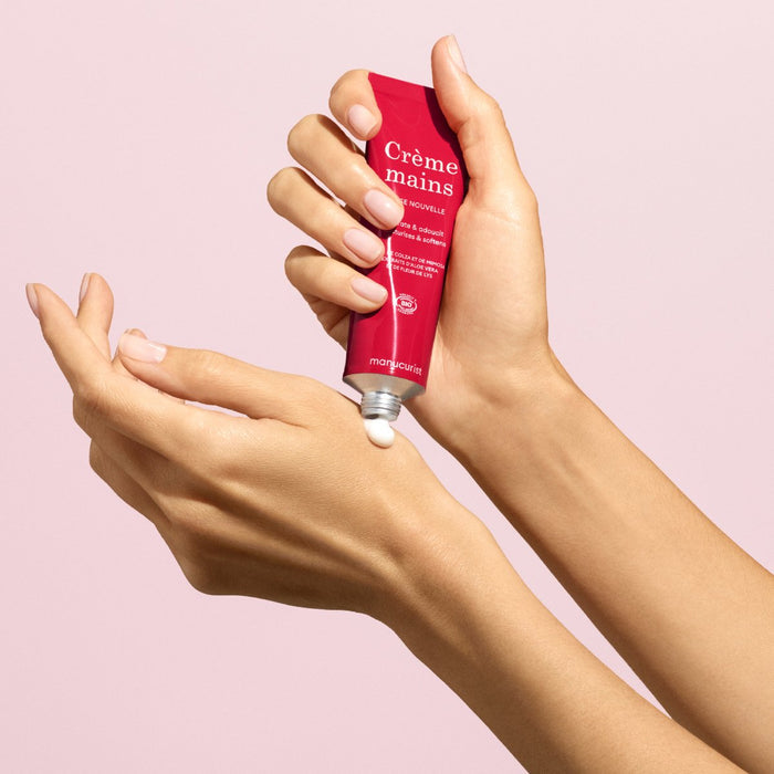 A person applies a small amount of Manucurist Everyday Rose Hand Cream - Cosmos Organic from a red tube onto the back of their hand, set against a light pink background.