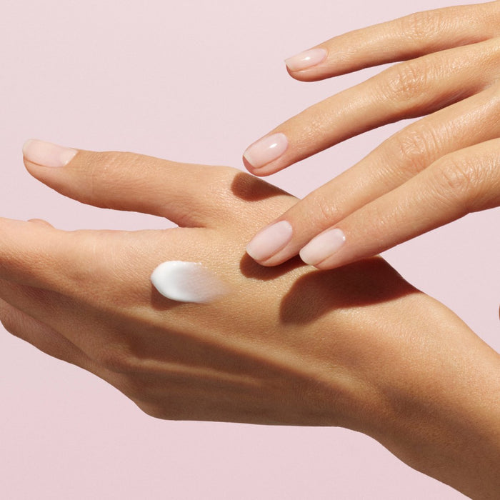 A close-up of a hand with manicured nails applying a small amount of Manucurist Everyday Rose Hand Cream - Cosmos Organic to the back of the other hand, set against a soft pink background.