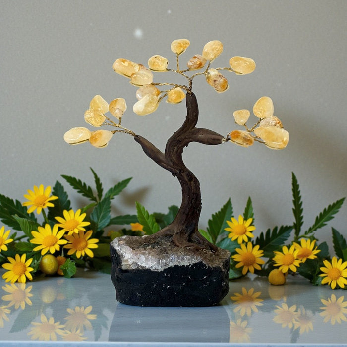 Decorative crystal tree with citrine  stones on a reflective surface with flowers in the background