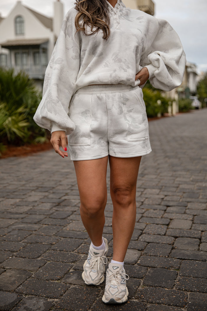 Person wearing a light gray outfit with shorts and sneakers on a paved walkway.