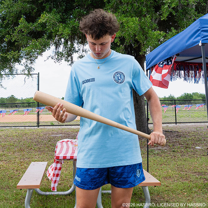 Adult male model wearing light blue GI Joe tee examining baseball bat at outdoor baseball event
