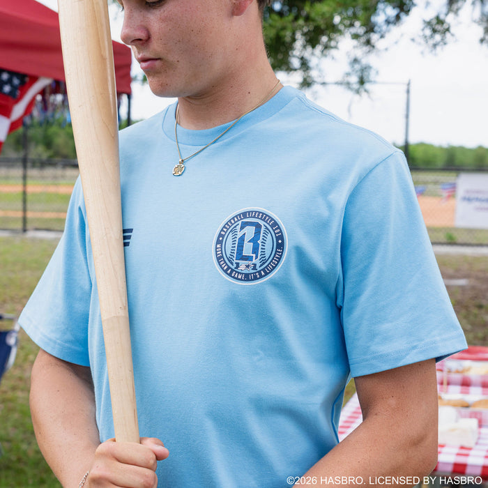 Blue: Close-up of adult male model in light blue GI Joe tee showing circular Baseball Lifestyle 101 badge while holding baseball bat