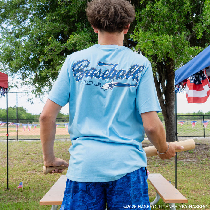Back view of adult male model showing Baseball Lifestyle 101 script graphic on light blue tee while holding a baseball bat at outdoor event