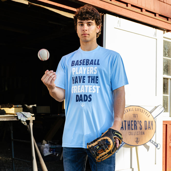 Young adult male model tossing a baseball while holding a fielding glove and wearing the columbia blue Greatest Dad Athletic Tee in front of a barn with the BL101 Father's Day Collection sign visible in the background.