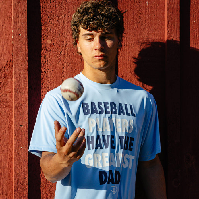 Young adult male model flipping a baseball into the air against a red barn wall while wearing the columbia blue Greatest Dad Athletic Tee with navy and white "Baseball Players Have the Greatest Dads" graphic on the front.