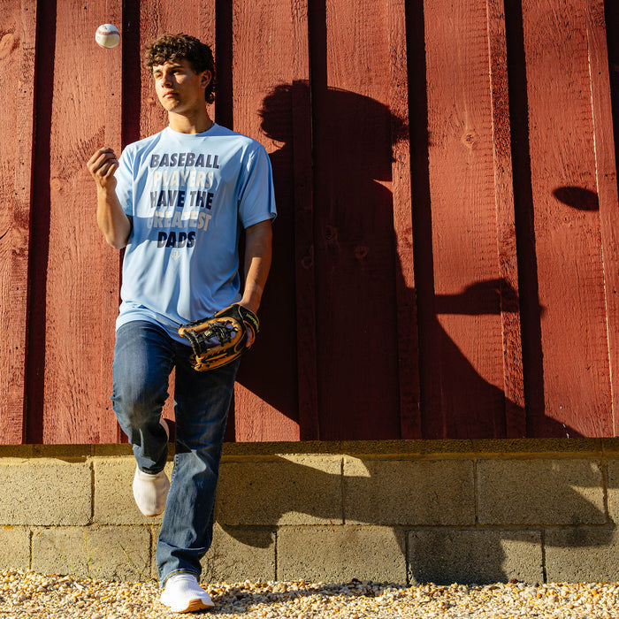 Young adult male model leaning against a red barn wall with a fielding glove and a baseball mid-air while wearing the columbia blue Greatest Dad Athletic Tee paired with jeans and white sneakers.
