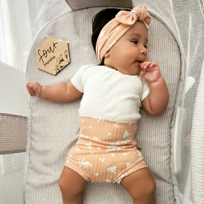 Baby in a crib wearing a white shirt and peach shorts with a headband, next to a 'four months' milestone card.