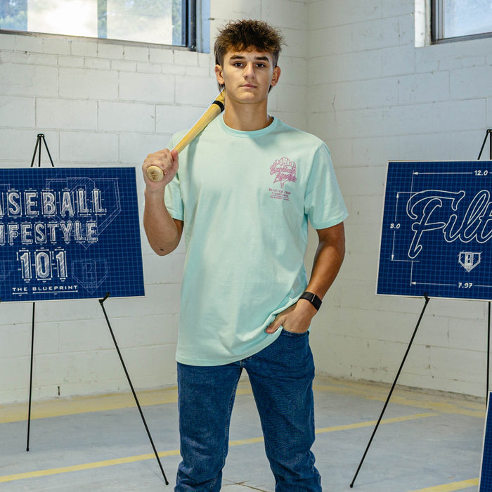 Person wearing light blue t-shirt with pink ice cream graphic and "Baseball Lifestyle 101" logo, while holding a baseball bat.