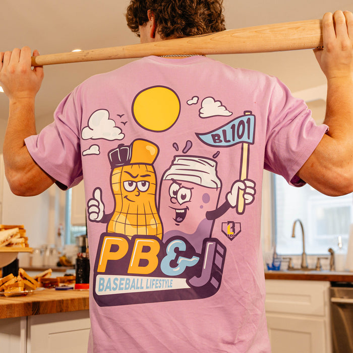 Back view of the BL101 PB&J Baseball Lifestyle purple tee on a male model holding a baseball bat, showing the cartoon peanut and jelly jar mascot graphic.