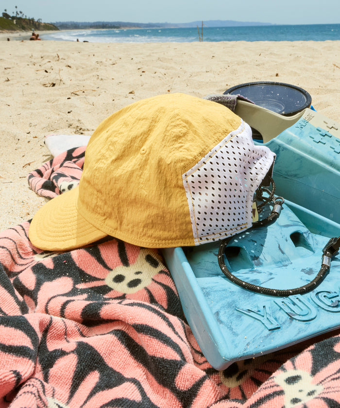 Yellow cap, sunglasses, and pink towel on a beach with a blue cooler.