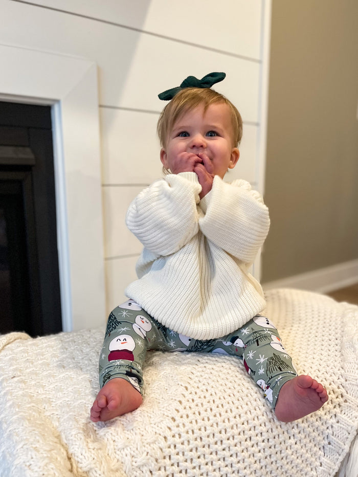 Baby sitting on a white surface wearing a large white bow and patterned pants.