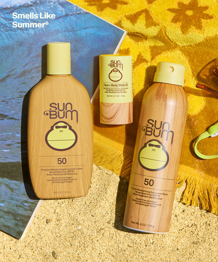 Sun Bum SPF 50 Spray + Lotion + Face and Body Stick 40 set on sand with yellow towel and beach accessories