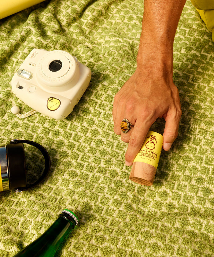 Hand holding Sun Bum SPF 50 Spray + Lotion + Face and Body Stick 40 on green blanket with camera and bottle nearby