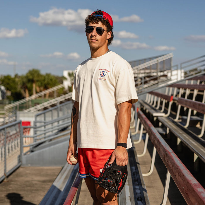 Male model wearing the BL101 America's Pastime cream tee and red mesh shorts at a baseball stadium, holding a baseball and glove.