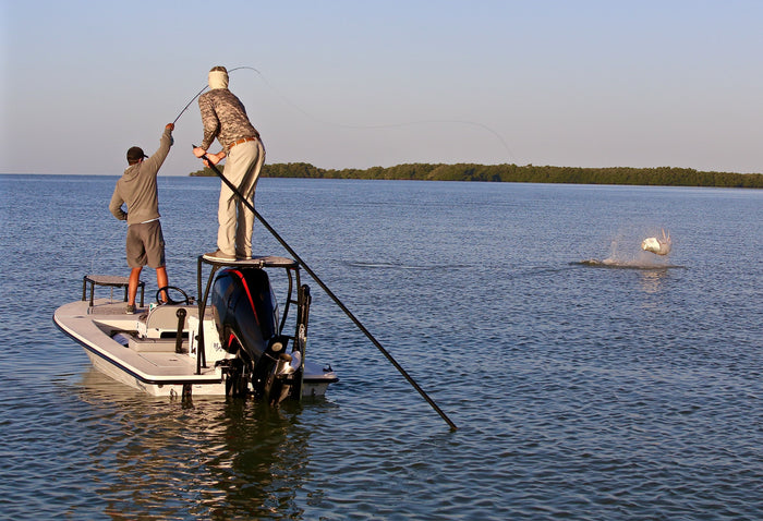 Two anglers fishing on a boat. One is holding onto his fishing pole, while a fish is jumping out of the water.