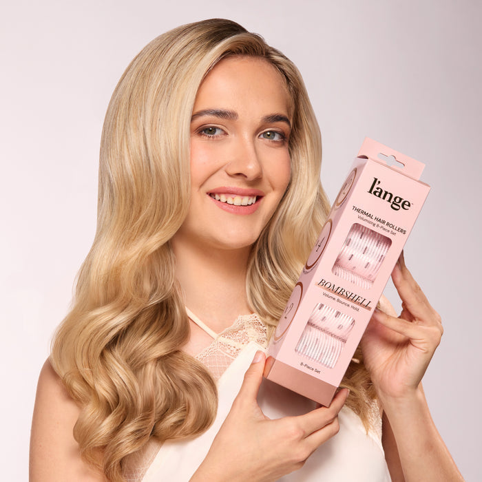 A smiling model with blonde hair holding a pink L’ange Thermal Hair Rollers box