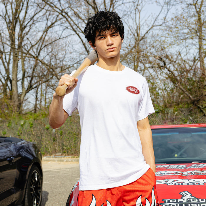 Young man holding baseball bat over shoulder, wearing white Throwing Gas tee with red BL101 chest oval, red flame shorts, beside red and black cars.