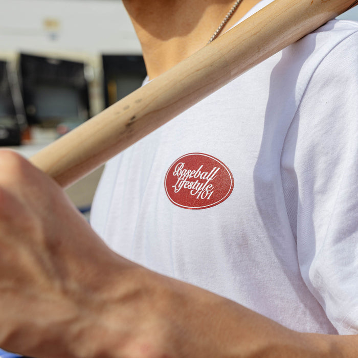 Close-up of red Baseball Lifestyle 101 oval chest graphic on white tee, wooden bat resting across chest.
