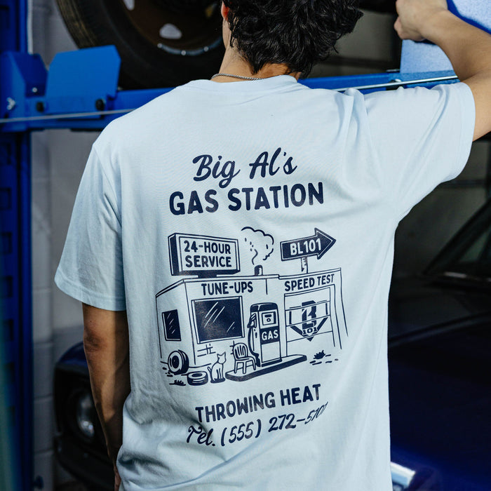 Back of light blue tee with navy vintage gas station illustration and "Throwing Heat" text, garage setting.