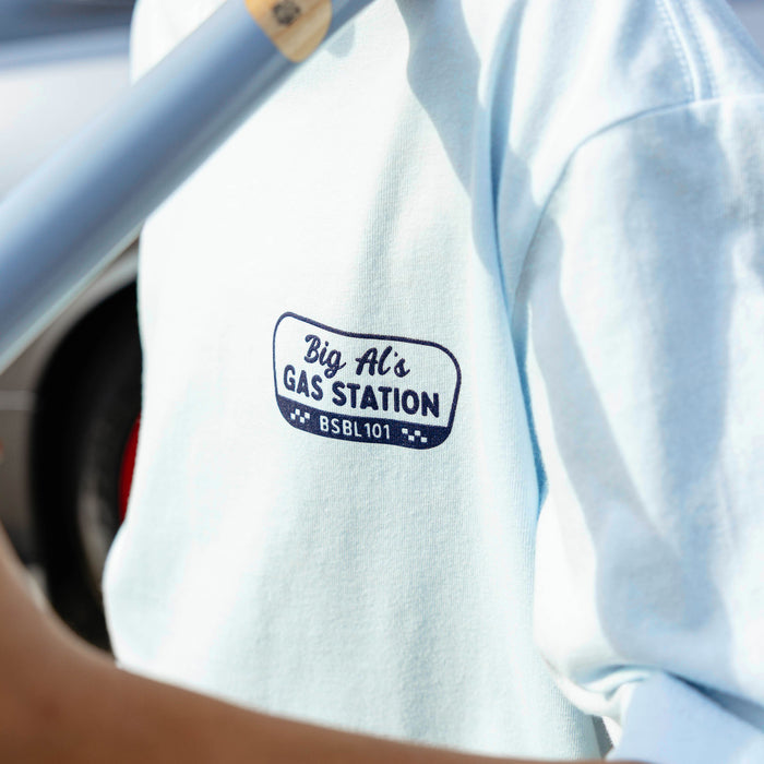 Close-up of navy Big Al's Gas Station BSBL 101 badge graphic on light blue tee, bat handle visible at top.