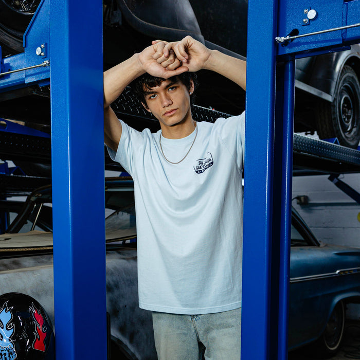Young man between blue automotive lift columns, arms raised, wearing light blue Big Al's Gas Station tee and light wash jeans.