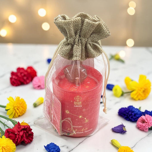 Aries  candle in a clear bag with a decorative bow, surrounded by colorful flowers on a light surface.
